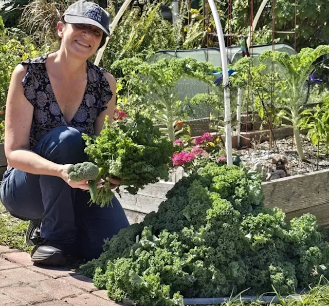 Melanie kneeling in the garden with a freshly harvested armful of broccoli and curly kale